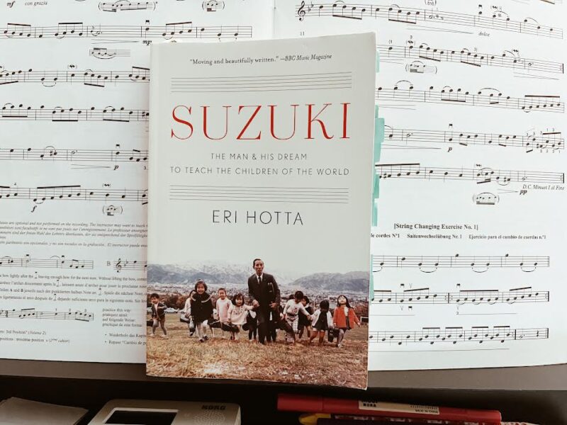 picture of Suzuki biography on a music stand in front of Suzuki Method books